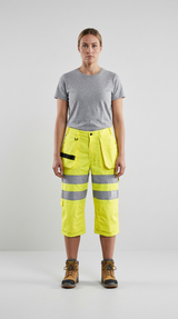 Kvinde i grå t-shirt, neon-gule 3/4 arbejdsbukser med reflekser og brune arbejdsstøvler.