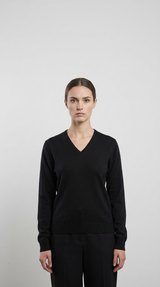 Kvinde iført en sort v-hals sweater og sorte bukser mod en lys grå baggrund.