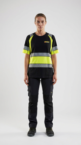Kvinde i sort og gul high-vis t-shirt med reflekser og mørke arbejdsbukser mod en lys baggrund.