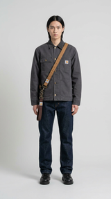 Mand i grå Carhartt-jakke, mørkeblå jeans og brun skulderrem foran en lys grå baggrund.