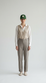 Kvinde i grøn kasket, hvid skjorte, beige vest og grå bukser mod en lys, neutral baggrund.