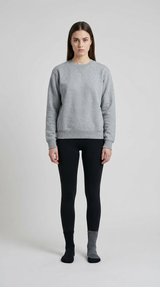 Kvinde i grå sweatshirt og sorte leggings stående mod en lys baggrund.