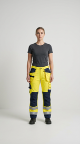 Kvinde i grå t-shirt og gule high-vis arbejdsbukser med reflekser mod en lys baggrund.