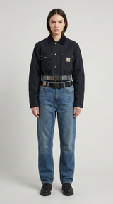 Kvinde i sort Carhartt-jakke og blå jeans med to bælter mod en grå baggrund.