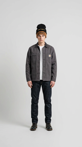 Mand i grå Carhartt-jakke, mørke jeans og to huer mod en lys grå baggrund.