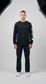 Mand iført sort langærmet Carhartt-trøje med logo på ærmet, mørke bukser og hvide sneakers i et fotostudie.