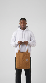 Ung mand i lysegrå hættetrøje holder en brun Carhartt-mulepose foran en neutral baggrund.