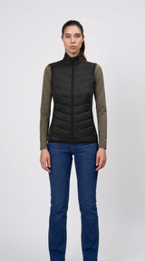 Kvinde i sort quiltet vest, olivengrøn langærmet trøje og blå jeans mod en hvid baggrund.