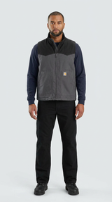 Mand i tofarvet grå Carhartt-vest, mørkeblå trøje og sorte bukser mod en lys baggrund.