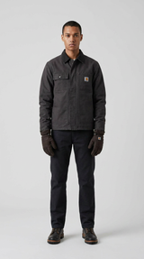 En mand i en mørk Carhartt-jakke, sorte bukser og brune handsker foran en grå baggrund.