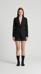 Kvinde i sort blazer og shorts med sorte strømper mod en lys grå baggrund.
