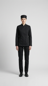 Kvinde i sort kokkeuniform med jakke, bukser og hue mod en lys grå baggrund.