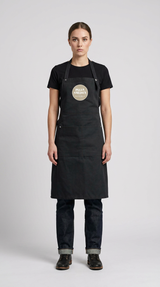 Kvinde i sort t-shirt og jeans iført et sort forklæde med 'Pizza Smeden'-logoet mod en grå baggrund.