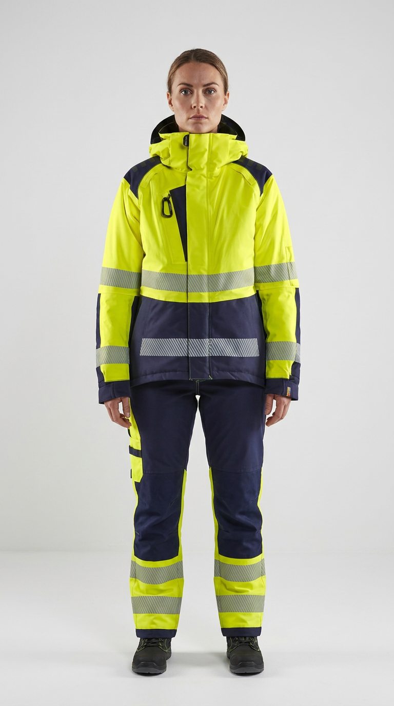Kvinde i gul og mørkeblå high-visibility arbejdsjakke og bukser med refleksstriber.