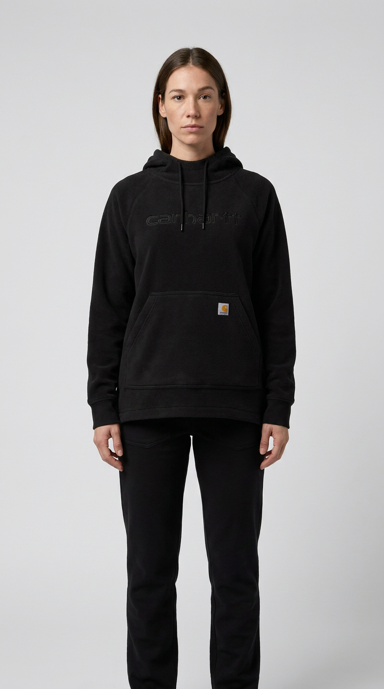 Kvinde i sort Carhartt fleece-hættetrøje og sorte bukser foran en grå baggrund.