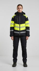 Kvinde i sort og neon-gul arbejdsjakke med refleksstriber og sorte arbejdsbukser på en grå baggrund.