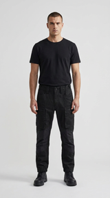 Mand i sort t-shirt og sorte cargobukser stående mod en hvid baggrund.