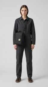 Kvinde i sort jumpsuit med en sort Carhartt skuldertaske mod en grå baggrund.