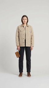Kvinde i beige Carhartt-jakke, mørke bukser og brune støvler, der holder en lille brun taske mod en grå baggrund.