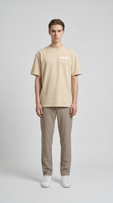 Mand i beige t-shirt med '10KCD' tekst, brune bukser og hvide sneakers mod en grå baggrund.