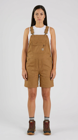 Kvinde i brune Carhartt-smækbukser, hvid tanktop og arbejdsstøvler mod en hvid baggrund.