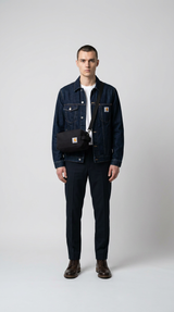Mand i denimjakke og mørke bukser med en sort Carhartt skuldertaske mod en hvid baggrund.