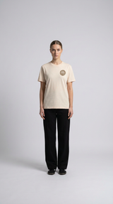 Kvinde i beige t-shirt med rundt logo og sorte bukser mod en lys grå baggrund.