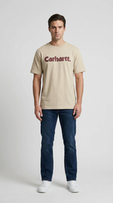 Mand i beige Carhartt t-shirt, mørkeblå jeans og hvide sneakers foran en grå baggrund.