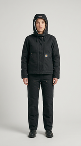 Kvinde iført sort Carhartt-jakke med hætte og matchende arbejdsbukser foran en grå baggrund.