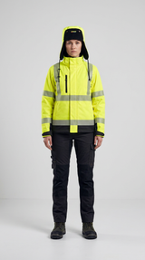 Kvinde i gul high-visibility arbejdsjakke, sorte arbejdsbukser og vinterhue foran en grå baggrund.