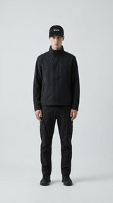 Mand i sort jakke, cargobukser og kasket med logo står foran en lys grå baggrund.