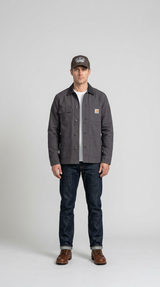 Mand iført grå Carhartt-jakke, mørkeblå jeans, brune støvler og kasket mod en neutral baggrund.