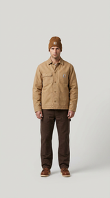 Mand i beige Carhartt-jakke, brun hue og mørke arbejdsbukser mod en lys grå baggrund.