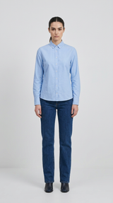 Kvinde i lyseblå langærmet skjorte og mørkeblå jeans mod en lys baggrund.