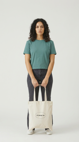 Kvinde i grøn t-shirt holder en cremefarvet mulepose med teksten "Dit design her" på en lys baggrund.