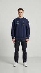 Mand iført mørkeblå sweatshirt med sponsorlogoer, mørke jeans og hvide sneakers mod en lys baggrund.