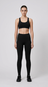 Kvinde i sort sports-bh og leggings stående mod en lys grå baggrund.