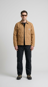 Mand iført en brun Carhartt-jakke, sorte bukser og solbriller mod en lys baggrund.