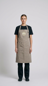 Kvinde i sort t-shirt og bukser iført et beige forklæde med "PIZZA SMEDEN" logo mod en hvid baggrund.