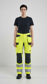 Kvinde i sort t-shirt og neon-gule arbejdsbukser med reflekser mod en lys baggrund.