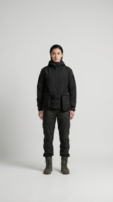 Kvinde i sort techwear-jakke, mørke cargobukser og støvler foran en lys grå baggrund.
