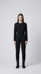 Kvinde i en langærmet sort jumpsuit og sorte snørestøvler mod en lysegrå baggrund.