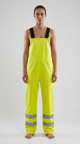 Kvinde i neon-gule overalls med sorte seler og refleksstriber på benene mod en lys baggrund.