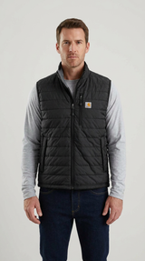 Mand iført en sort quiltet Carhartt-vest, grå langærmet trøje og mørkeblå jeans mod en lys baggrund.
