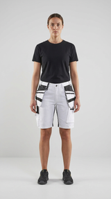 Kvinde iført sort t-shirt og hvide arbejdsshorts med hængelommer foran en grå baggrund.