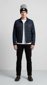 Mand i mørkeblå denimjakke, hvid t-shirt, sorte bukser og hue stående mod en grå baggrund.