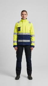 Kvinde i gul og marineblå high-visibility arbejdsjakke og sorte arbejdsbukser mod en grå baggrund.