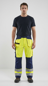 Mand i sort t-shirt og gul og blå high-visibility arbejdsbukser med refleksbånd på en lys baggrund.