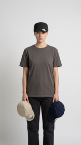 Kvinde i grå t-shirt og sort kasket holder en beige og en mørkeblå kasket mod en hvid baggrund.
