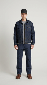 Mand i mørkeblå denimjakke, sort kasket og mørke bukser mod en lys grå baggrund.